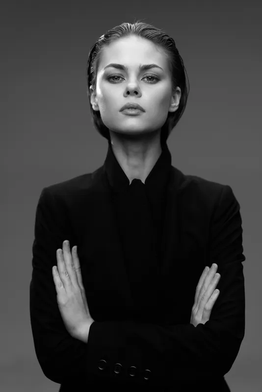 Black and white portrait of confident woman with arms crossed wearing dark jacket, serious expression on gray background in studio.