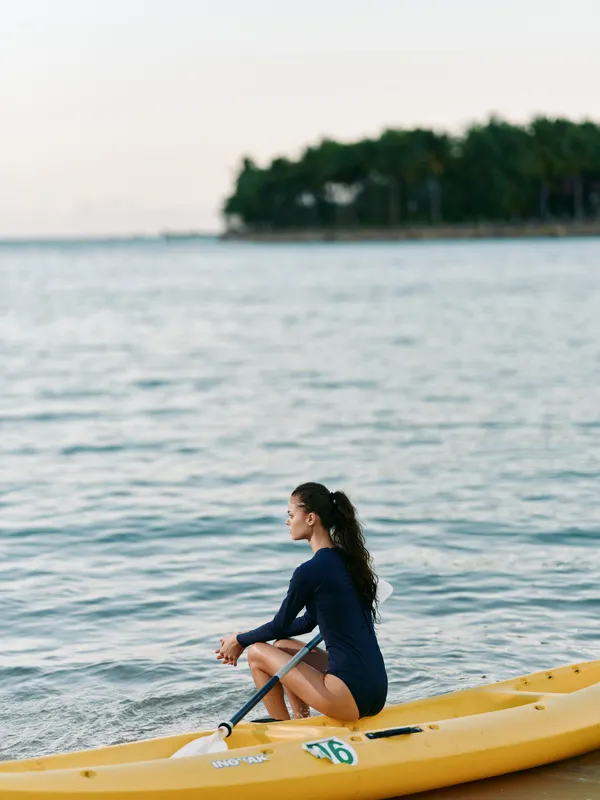 A young woman sits in a yellow kayak on calm water near a quiet shoreline, paddling with focus as distant trees line the horizon under soft evening light.