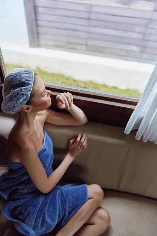 Relaxed woman in blue towel and shower cap sitting indoors by window, enjoying natural light and calm moment in cozy home setting with soft neutral tones.