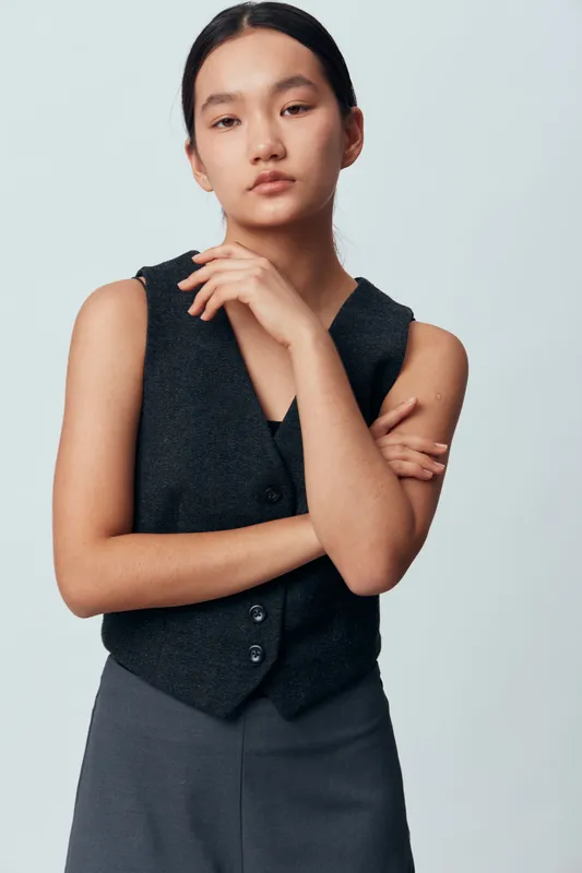 Young woman with straight dark hair wearing sleeveless dark gray vest and gray pants standing with arms crossed against neutral background in studio portrait.