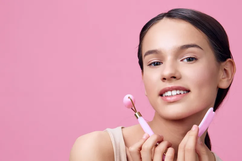 Young model with a bright smile holding pink makeup tools against a soft pink background, showcasing beauty and skincare concepts for modern aesthetics