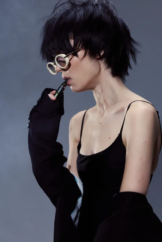 Stylish woman with short hair and glasses smoking vape in black outfit against gray background