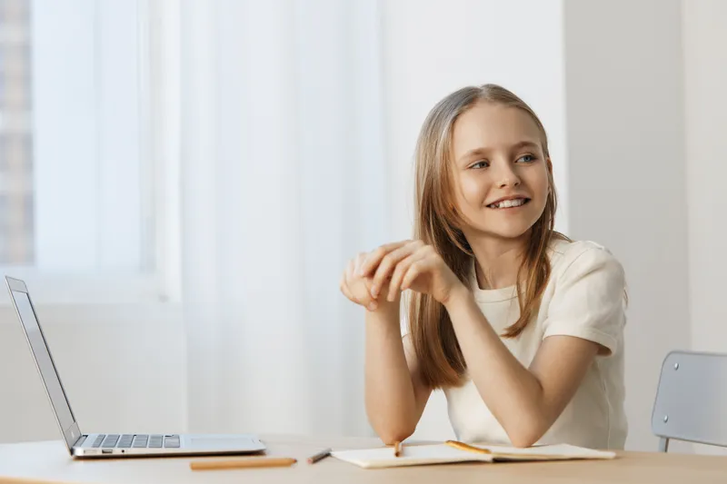 Cheerful young girl with long hair smiling while studying at a desk with a laptop, representing a positive learning atmosphere in a light colored room