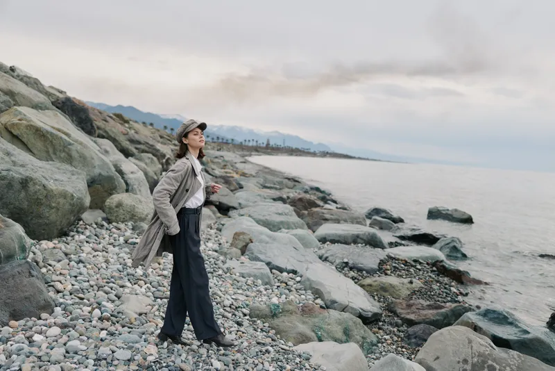 Young woman in casual outfit stands on rocky shore looking at sea under cloudy sky. Outdoor lifestyle, nature exploration, and peaceful coastal atmosphere concept.