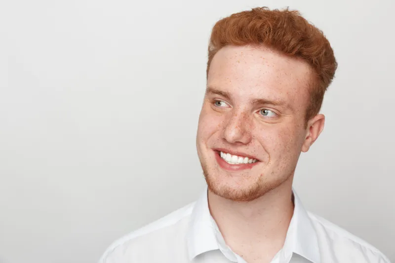 Smiling young redhead man with freckles looks away wearing white shirt on light background. Concept of happiness, youth, casual style, and positive emotions.