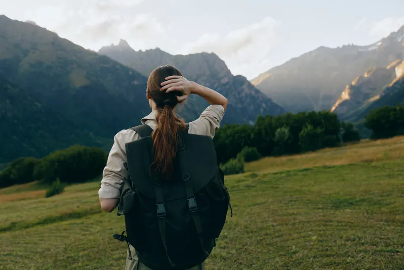 Woman with backpack standing in a green field looking at mountain range during daytime. Concept of lifestyle, adventure, and outdoor exploration in nature environment.