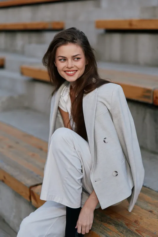 Confident business woman in a modern outfit, sitting on wooden steps, smiling brightly, showcasing a contemporary style and relaxed attitude.