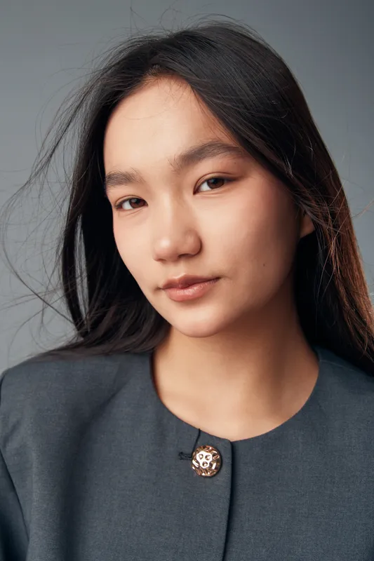 Young woman with long black hair and natural makeup wearing gray blazer with decorative button looking at camera with calm expression on plain white background closeup portrait.