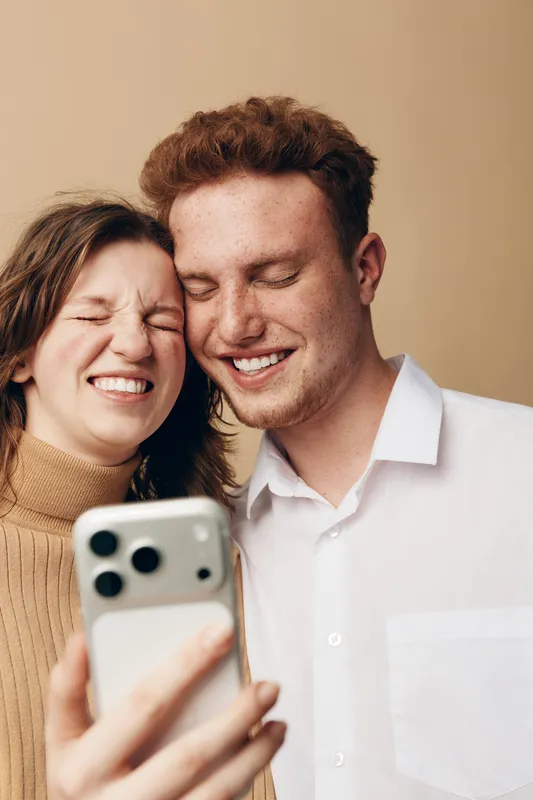 Happy young couple with freckles smiling and taking a selfie together on smartphone. Casual clothing and warm beige background create a cozy and joyful atmosphere of friendship and love.