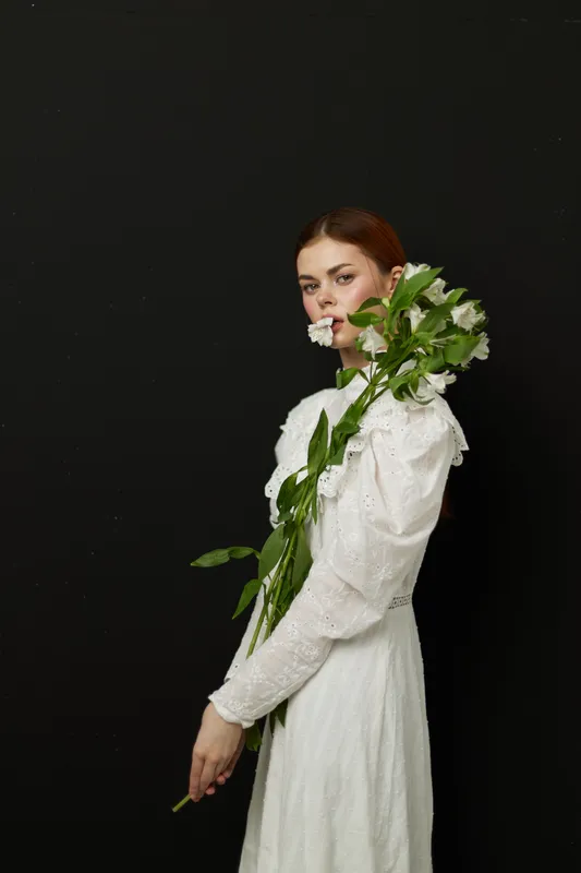 Elegant young woman in a white dress holding flowers against a black background, expressing tranquility and beauty, perfect for spring themes and aesthetics