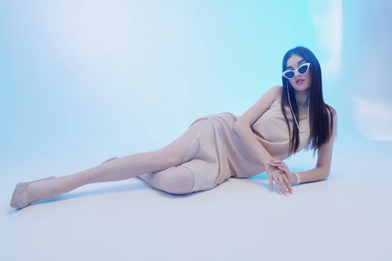 Fashionable woman with long hair wearing beige dress and white sunglasses lies on the floor against a light blue background. Concept of beauty, fashion, and style.