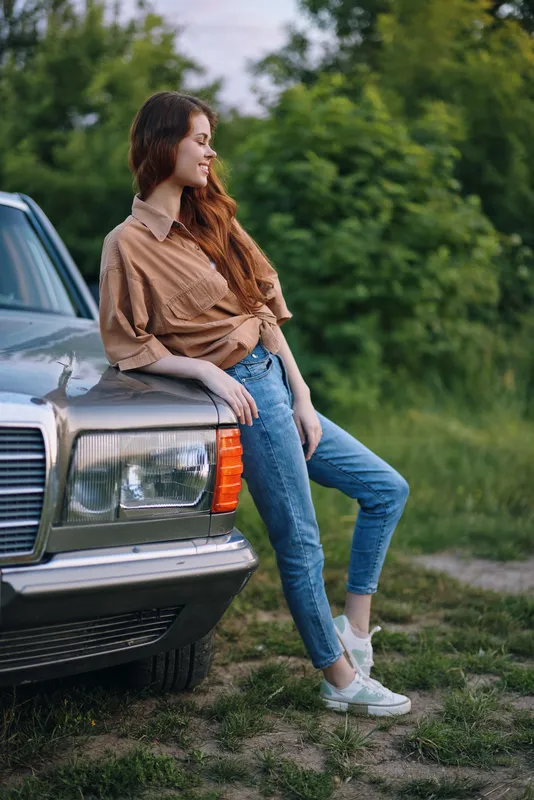 Young woman in casual attire leaning on a vintage car, showcasing a relaxed summer mood amidst greenery, evoking feelings of freedom and adventure.
