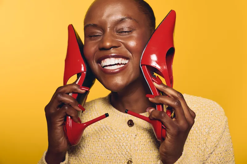 Happy African woman with dark skin holds red high heels near face smiling with closed eyes on yellow background. Concept of fashion, beauty, joy, and stylish accessories.