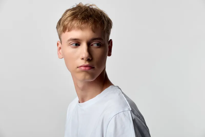 Teen boy looking aside wearing a white shirt in a neutral studio, conveying calm, curiosity, and focus. Simple lighting highlights clean style and natural expression, suitable for fashion, youth,