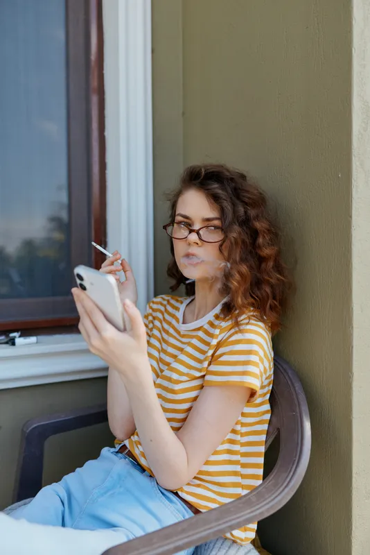 A young woman with curly hair and glasses sits on a porch, scrolling a smartphone, wearing a striped yellow tee and light shorts, capturing a quiet, everyday moment outdoors.