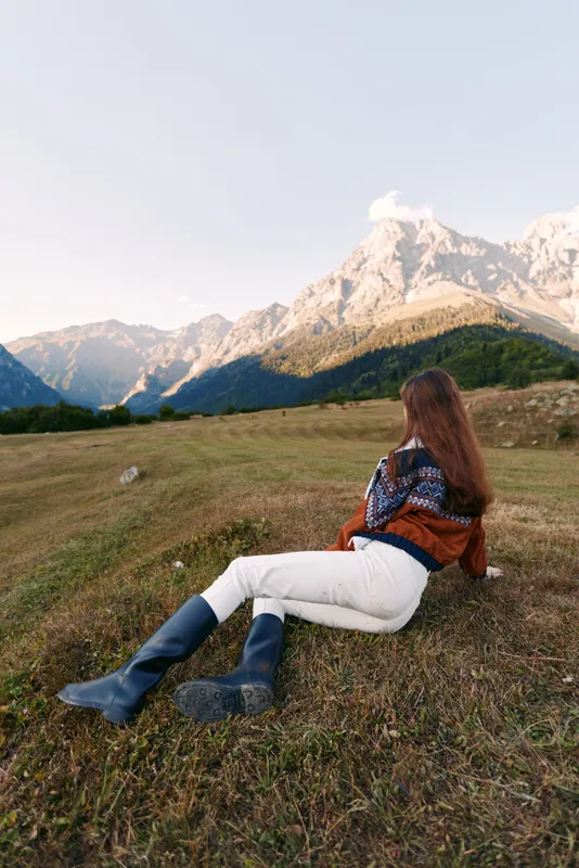 Woman lying on meadow facing mountain landscape, enjoying nature and wearing boots and sweater. Scenic outdoors view, peaceful relaxation and hiking retreat in open field.