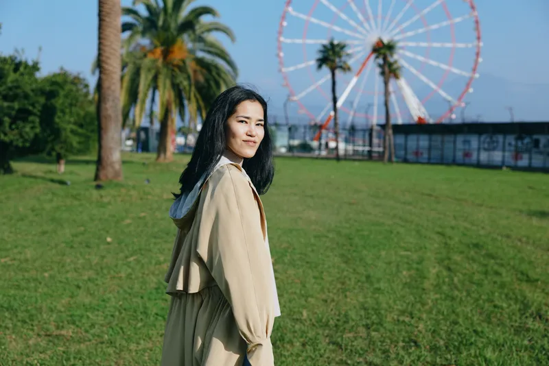 Woman in a beige coat in a sunny park beside a ferris wheel, casual outdoor scene with open space, calm expression, travel vibe and leisure photography