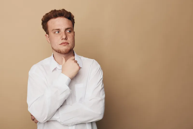 Thoughtful young man with red hair and freckles wearing white shirt looks away with hand on chin. Neutral beige background creates calm mood. Concept of contemplation and decision making.