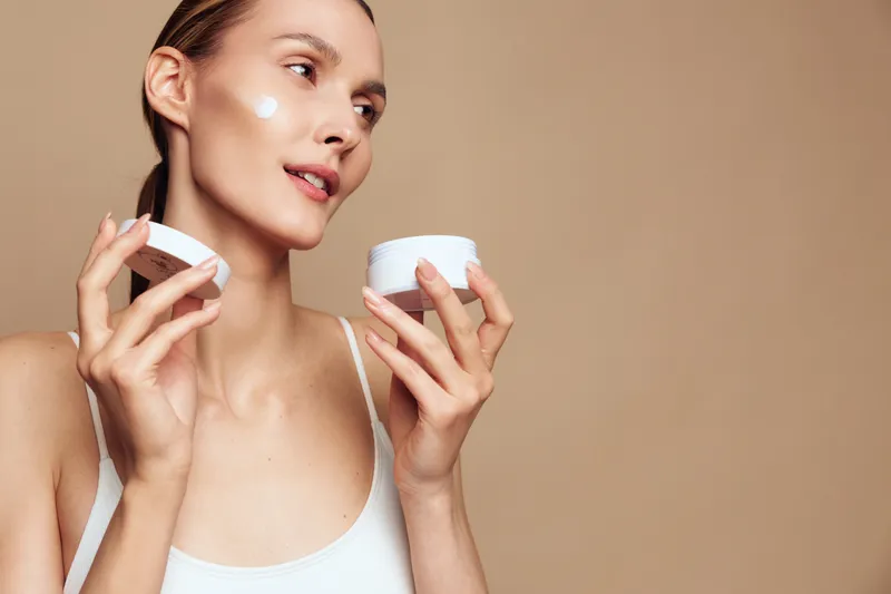 A young woman in a white tank top applies face cream on her cheek while holding an open jar against a beige background. Concept for skincare, beauty, and self-care routine.