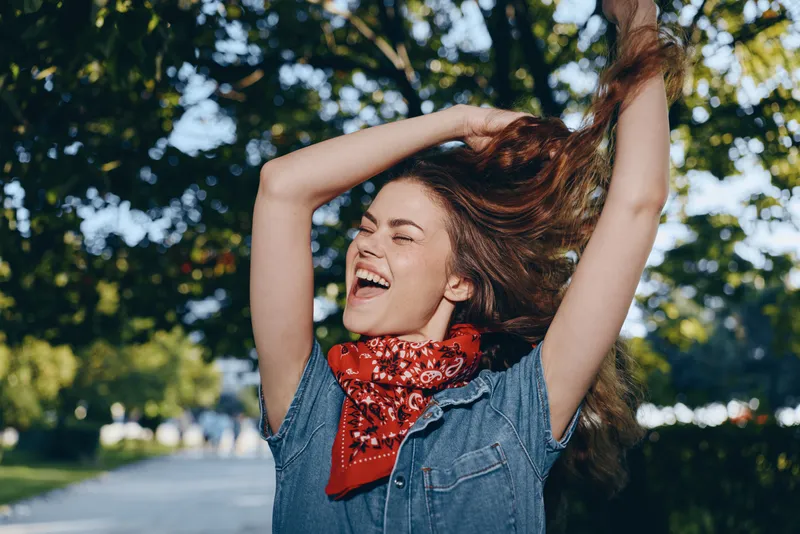 happy young woman with long hair and red bandana enjoying sunny day outdoors in green park with arms raised joyfully