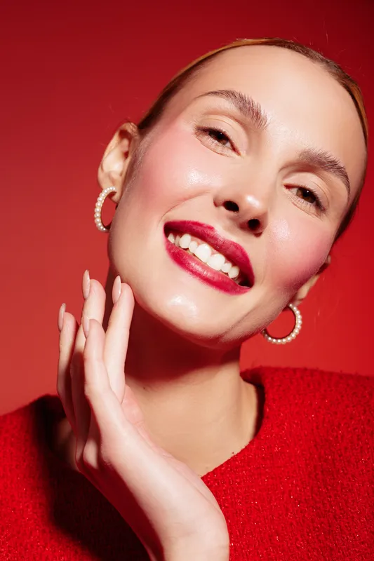 Smiling woman with red lipstick and hoop earrings poses against red background. Close-up portrait with glowing skin and joyful expression. Concept of beauty, fashion, and confidence.