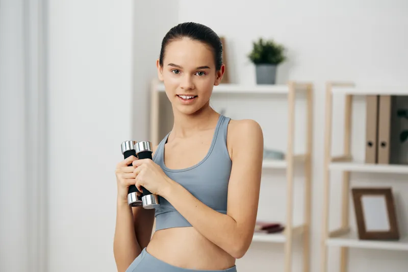 Woman in athletic wear lifting dumbbells at home, showcasing fitness and training routine with focus, balance and strength gains in a bright, clean room.