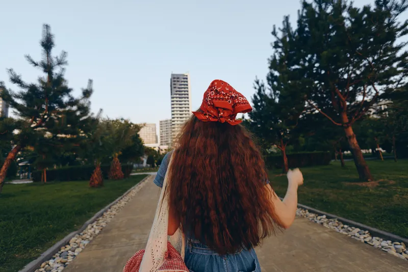 lifestyle woman in modern boho-western denim street style walking outdoors, film color with long curly hair and red bandana on urban pathway, casual and trendy outfit