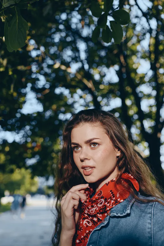 lifestyle woman in modern boho-western denim street style with a red bandana captured in warm film color tones outdoors under green trees