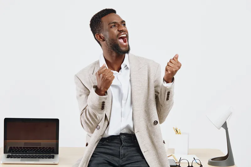 Smiling young man in casual office attire expressing happiness and excitement, standing at desk with laptop and stationery, isolated on plain white background. People, celebration, success concept
