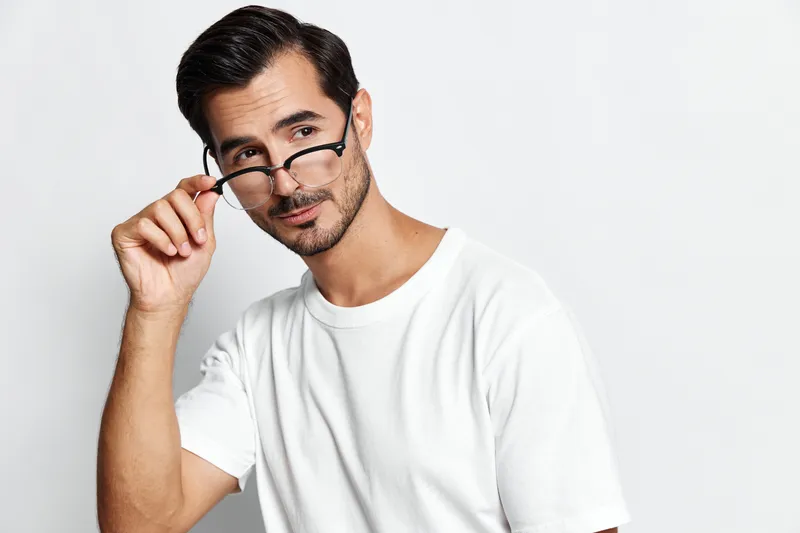Handsome confident young man adjusting glasses while looking at camera, wearing a casual white t-shirt, isolated on plain light grey background. Style and personality concept.