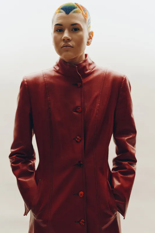 young woman with colorful short hair wearing red leather jacket stands against plain background, featuring modern urban style with confident expression and minimalistic composition.
