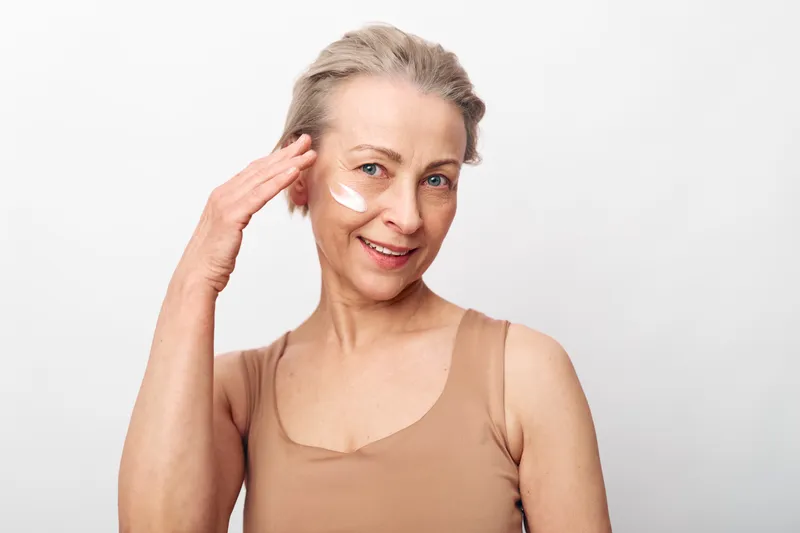 Smiling mature woman applies cream on face with fingers, wearing beige tank top. Concept of skincare, healthy aging, natural beauty, and self-care in bright studio setting.
