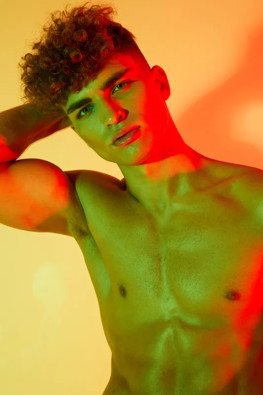 Young man with curly hair posing shirtless under colorful red and green lighting, showing muscular upper body and confident expression in studio portrait photography.