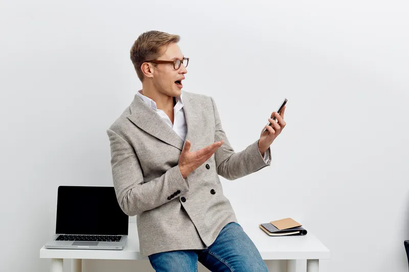 surprised man holding smartphone wearing glasses modern jacket sitting near laptop digital nomad concept remote work technology communication