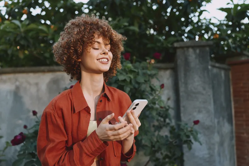 Smiling woman with curly hair using a smartphone outdoors in a garden, casual attire, natural light, lifestyle moment, warm, confident mood, everyday connection
