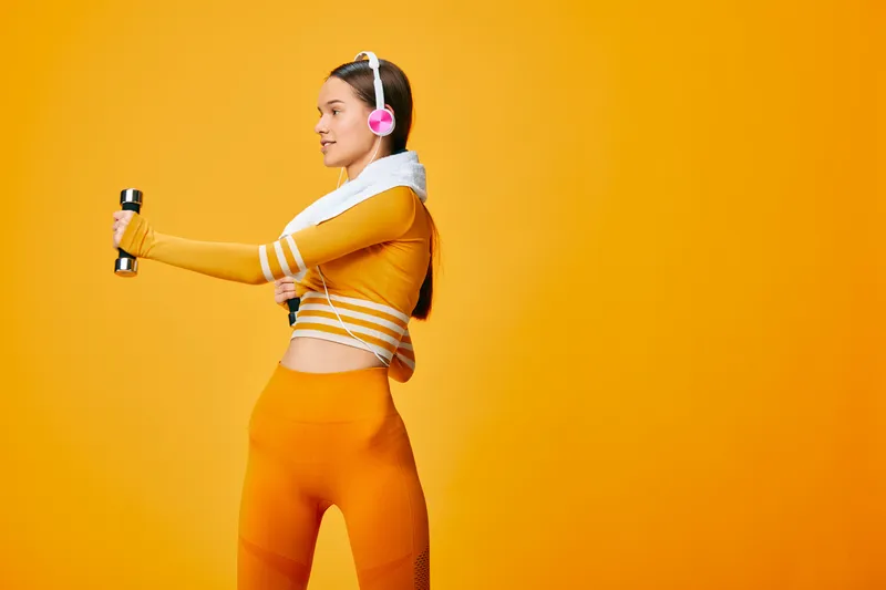 Fitness focused woman in bright orange athletic wear lifts dumbbells in a vivid orange studio, wearing pink headphones, showcasing energetic training and body movement.