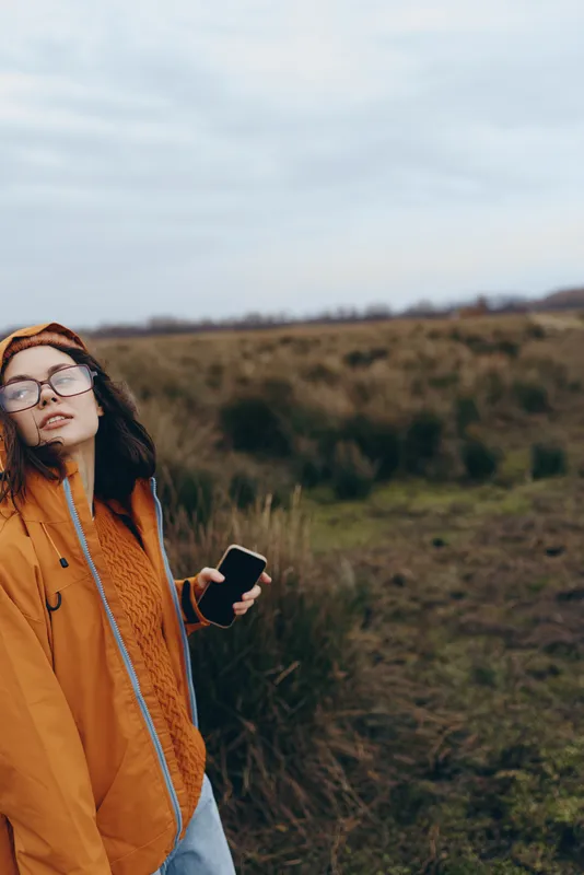 Woman lifestyle in an outdoor grassy field, orange jacket and glasses, holding a smartphone, calm expression, communication while walking in countryside landscape, cloudy sky today