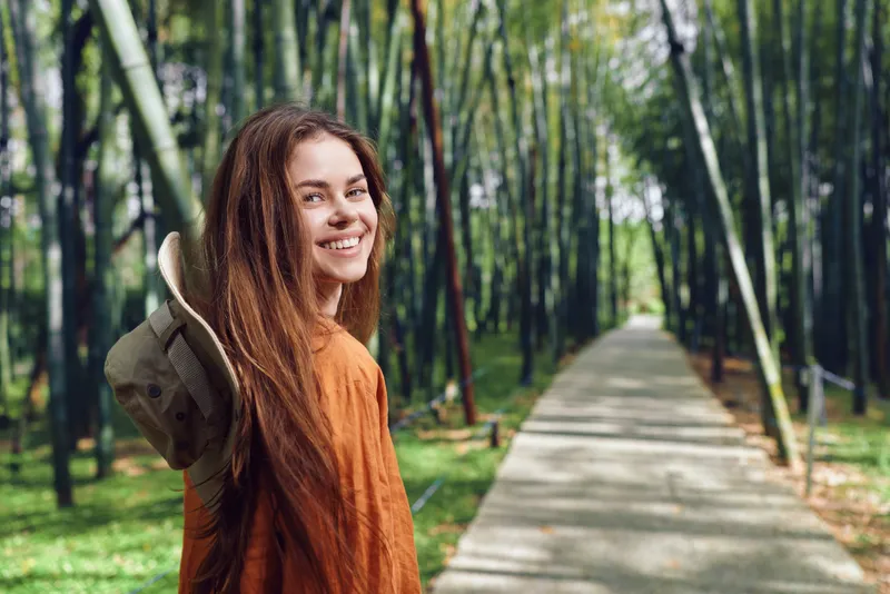 Woman smile backpack forest path nature travel hiking portrait of a young woman with long hair looking back on a scenic bamboo walkway, happy and relaxed outdoors in sunlight.