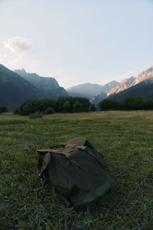 A lone backpack rests on a grassy meadow with distant mountains, conveying outdoor travel, hiking, and nature exploration in a serene landscape. Inviting solitude.
