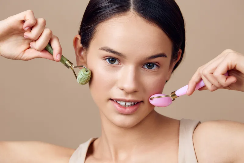 Young woman demonstrating facial massage techniques with jade and rose quartz rollers, promoting healthy skin and wellness in a light beige background