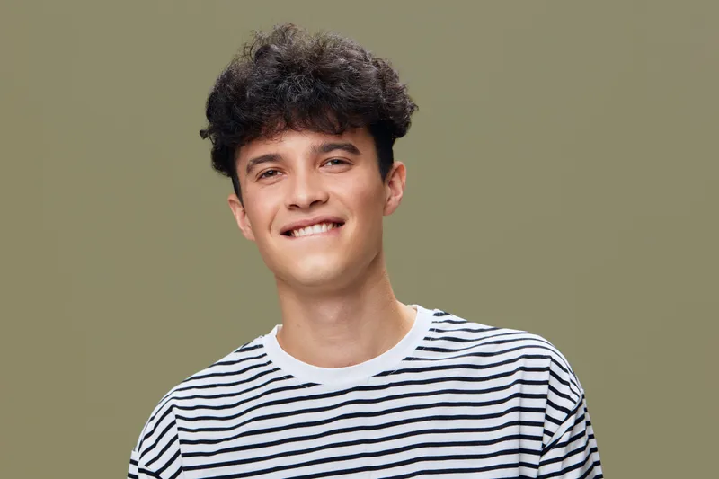 Young man smiling confidently in a casual striped shirt against a plain olive background, displaying relaxed and friendly expression.