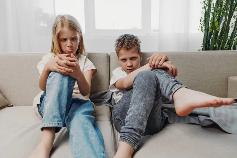 Two upset children sitting on the couch with crossed arms and serious expressions in a bright living room. Concept of sibling conflict, emotions, and family tension.
