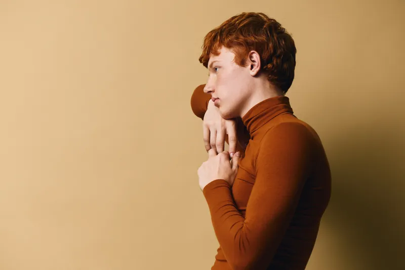 Young person with red hair and fair skin in brown turtleneck looking thoughtful in profile on beige background for portrait and contemplation.