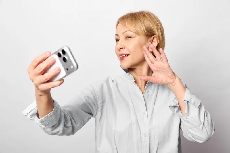 Senior woman holding smartphone, calling communication gesture near ear, worried face shows stress, indoor studio lighting, close up concept for mental health