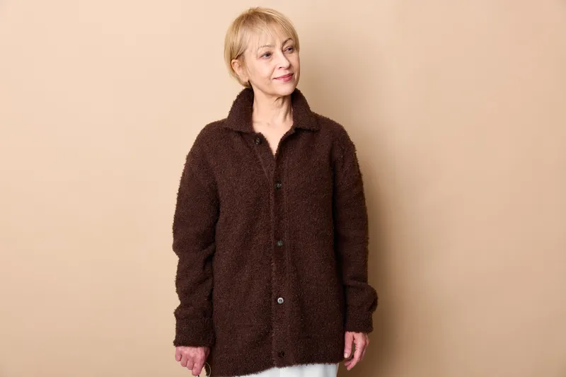 Woman portrait indoors beige background, senior brown jacket coat, standing pose, calm face, soft studio lighting, neutral mood, home interior feeling, mature age