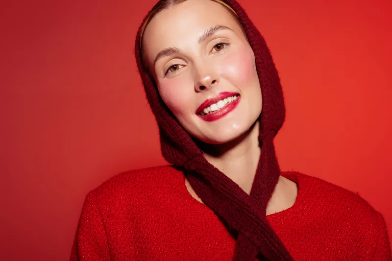 Smiling woman in red sweater and scarf poses against red background. Close-up portrait with bright makeup and glossy lips. Concept of fashion, beauty, and winter style.