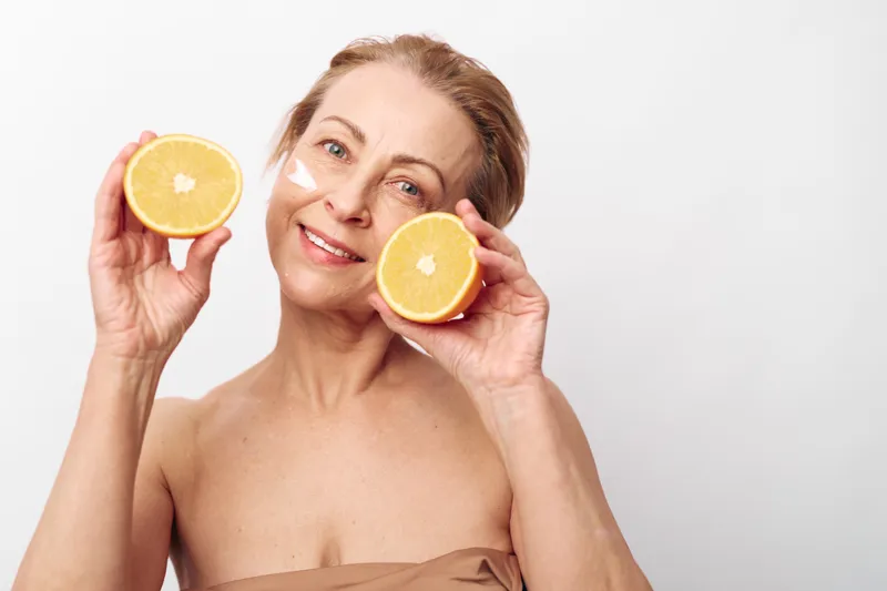 Mature woman with glowing skin holds two orange halves near face, applying cream under eyes. Concept for natural skincare, healthy aging, wellness, and beauty routine on white background.
