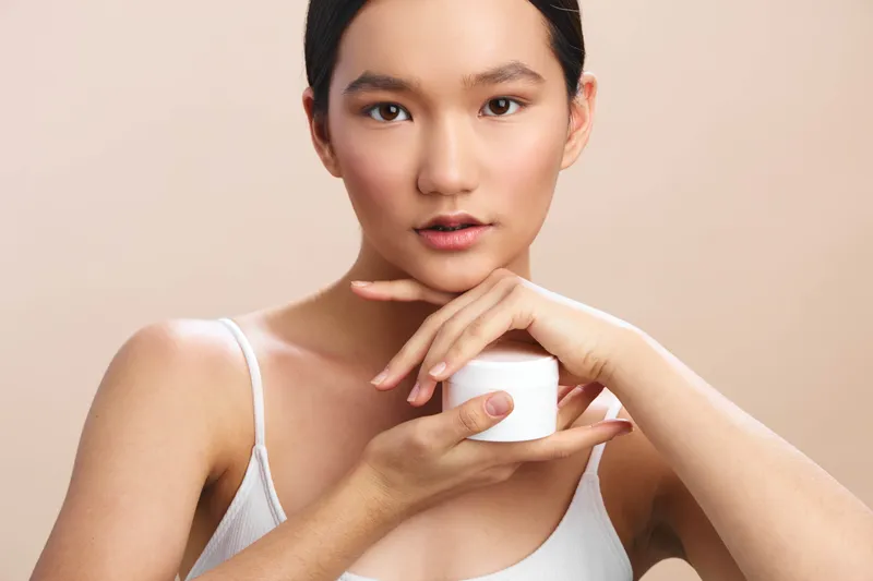 Asian woman holds a small cream jar near her face, looking softly at the camera with a calm smile, showcasing a skincare routine in a clean studio setting.