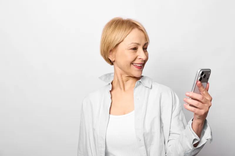 Woman senior smartphone smiling indoors communication technology portrait, white studio background, relaxed posture, short hair, holding phone, friendly expression, bright light, copy space