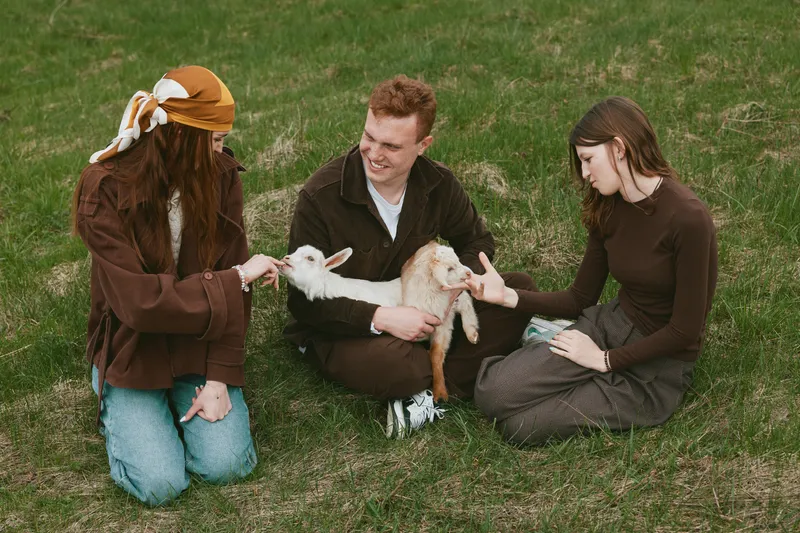 A relaxed group sits on grass, interacting with a cheerful goat kid, smiles and gentle touches, casual clothing, sunny outdoor scene. A wholesome moment of companionship between people and animals in